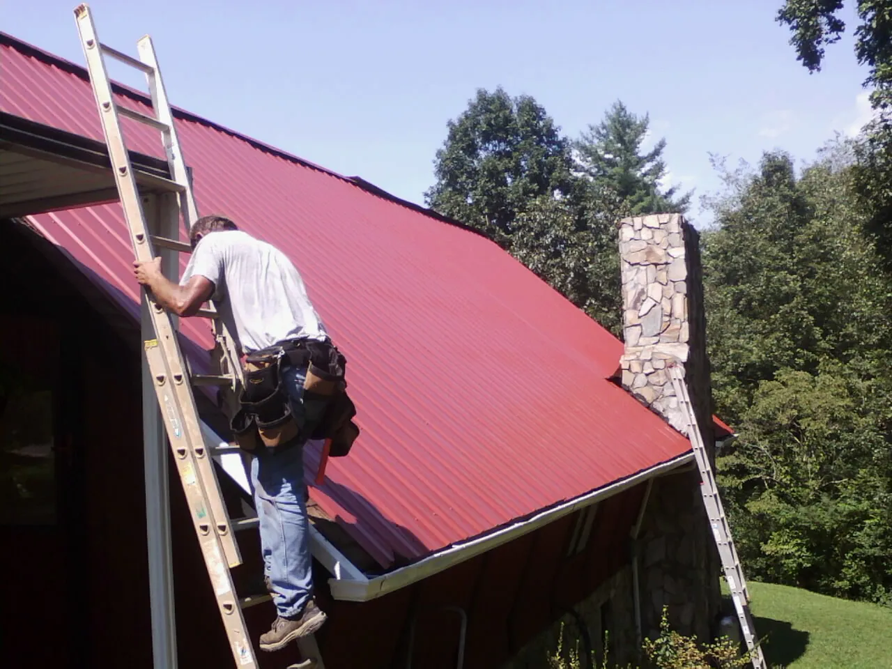 Roofer on a ladder beside a red metal roof and stone chimney — Soffit & Fascia Repair by Roofing Brothers in Kansas City